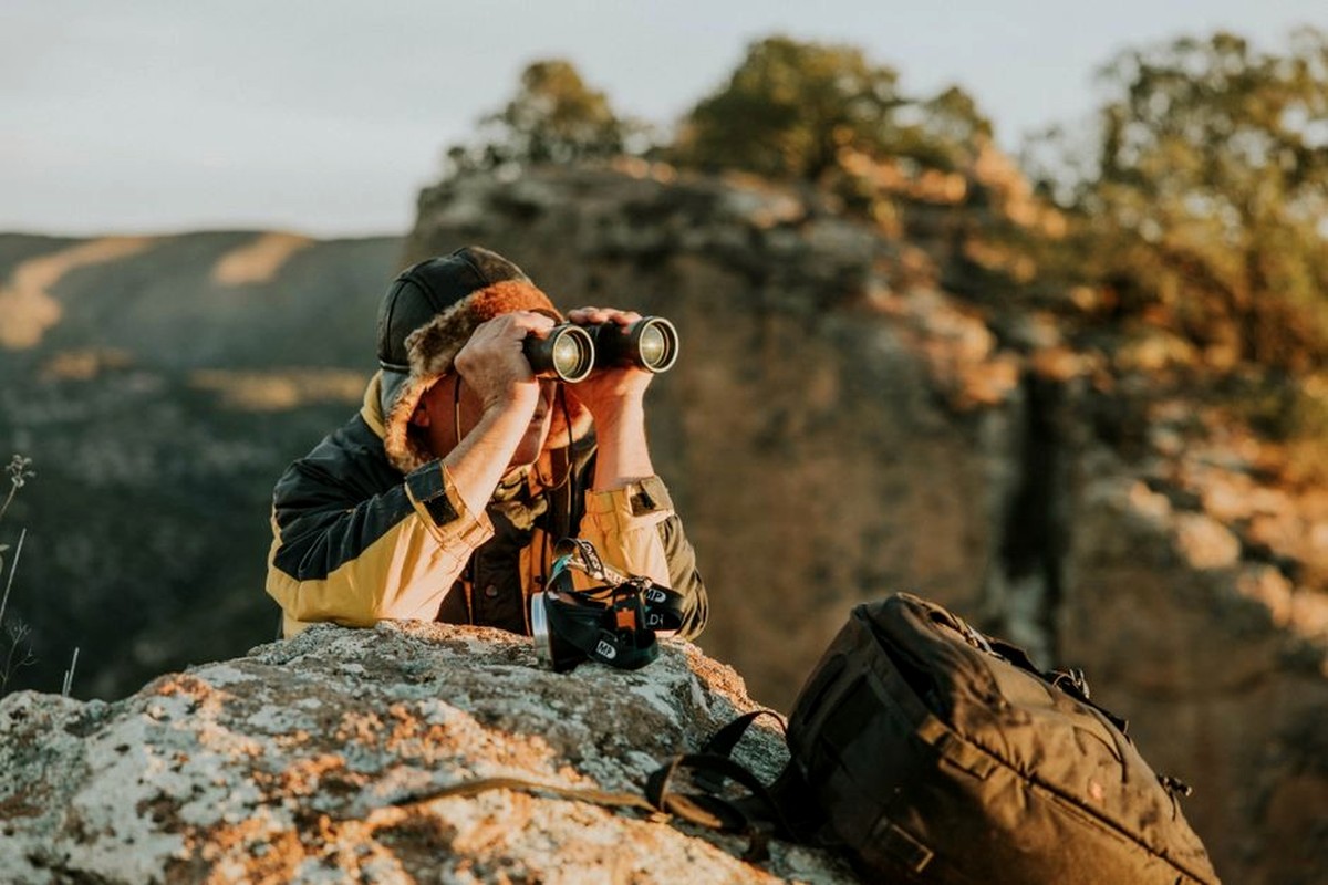 Outdoor observer on a rocky overlook with binoculars and a camera.